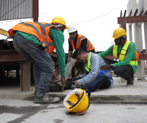 Construction workers in high-visibility vests and hard hats crouch around an injured person, providing first aid while one worker steadies a bright yellow helmet on the ground.