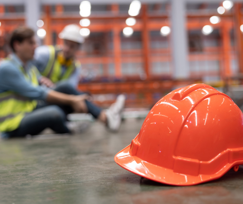 An orange hard hat lies on a concrete floor in a brightly lit warehouse, with workers in high-visibility vests sitting and working in the blurred background.